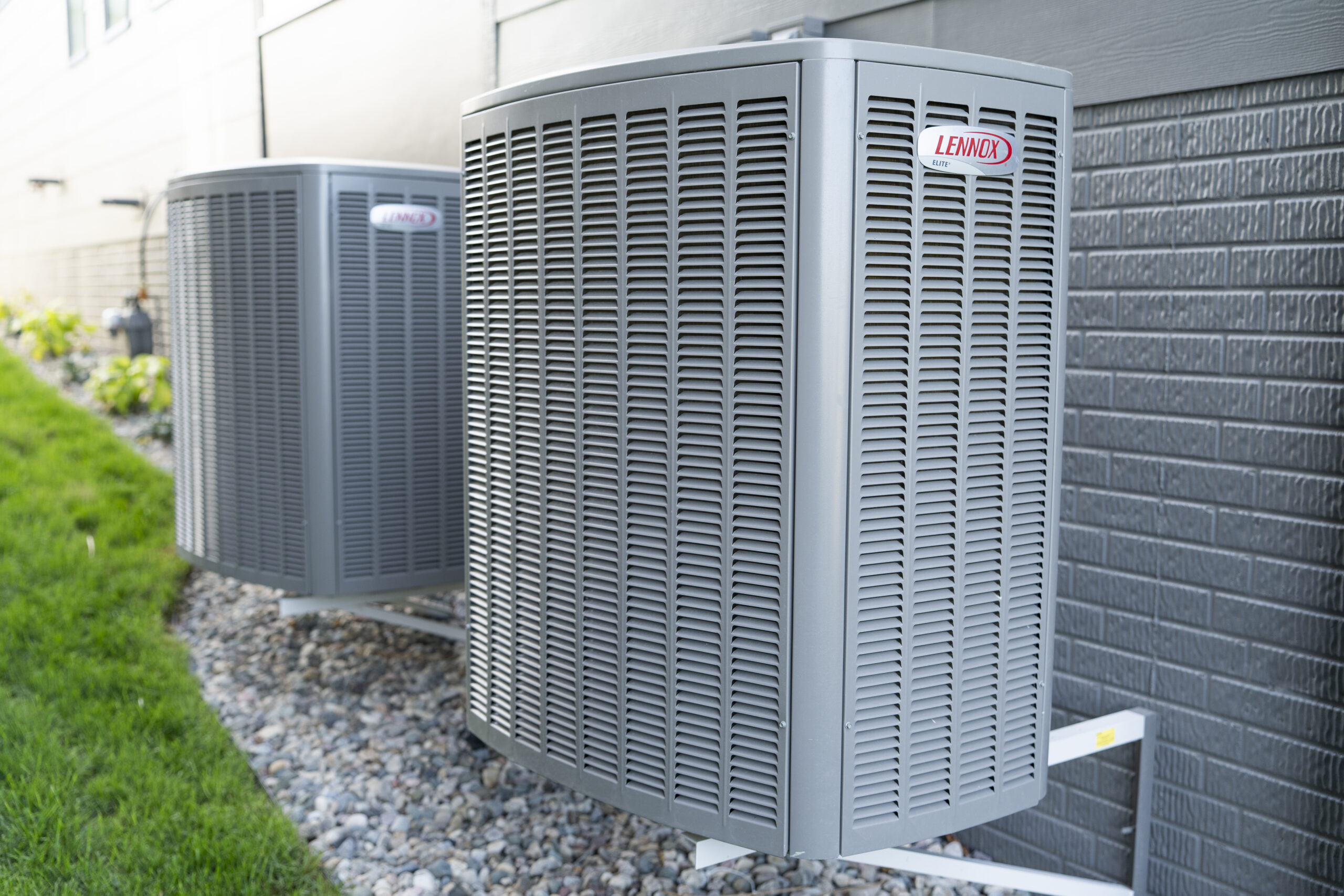 Two Lennox Elite HVAC condenser units mounted on an exterior wall over a pebble landscape.